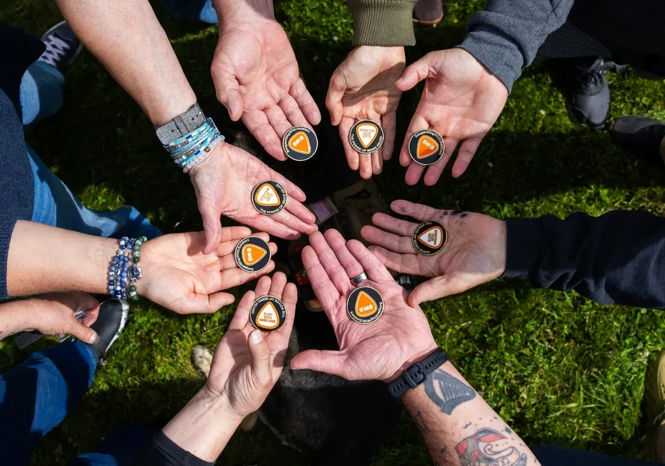 A circle of veterans’ open hands, each holding the same guitar-pick challenge coin